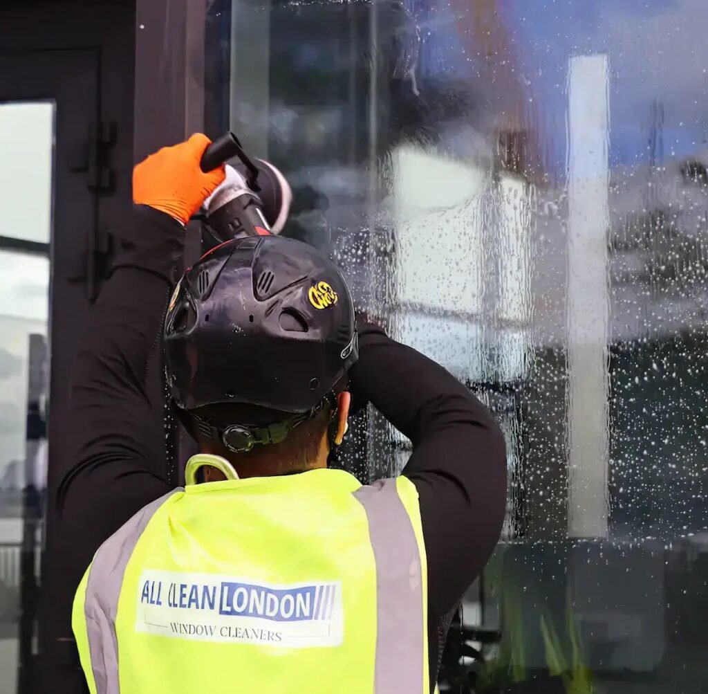 Window cleaner in a yellow safety vest and helmet scrubs a glass storefront with a power tool, water droplets on the glass left to right.